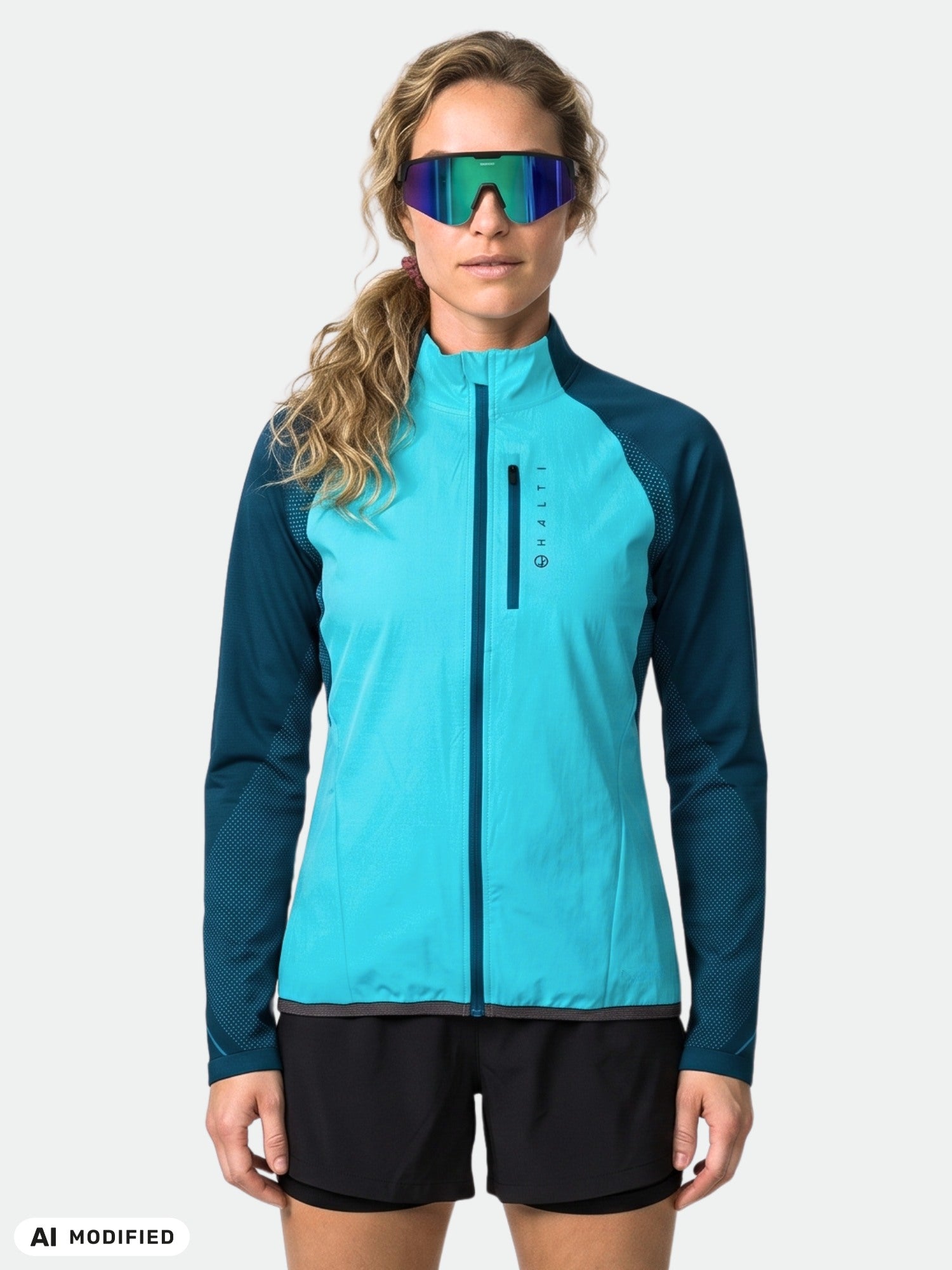 Model in Halti Tracks Seamless Jacket Women's (blue) with black shorts and reflective sunglasses, against plain backdrop.