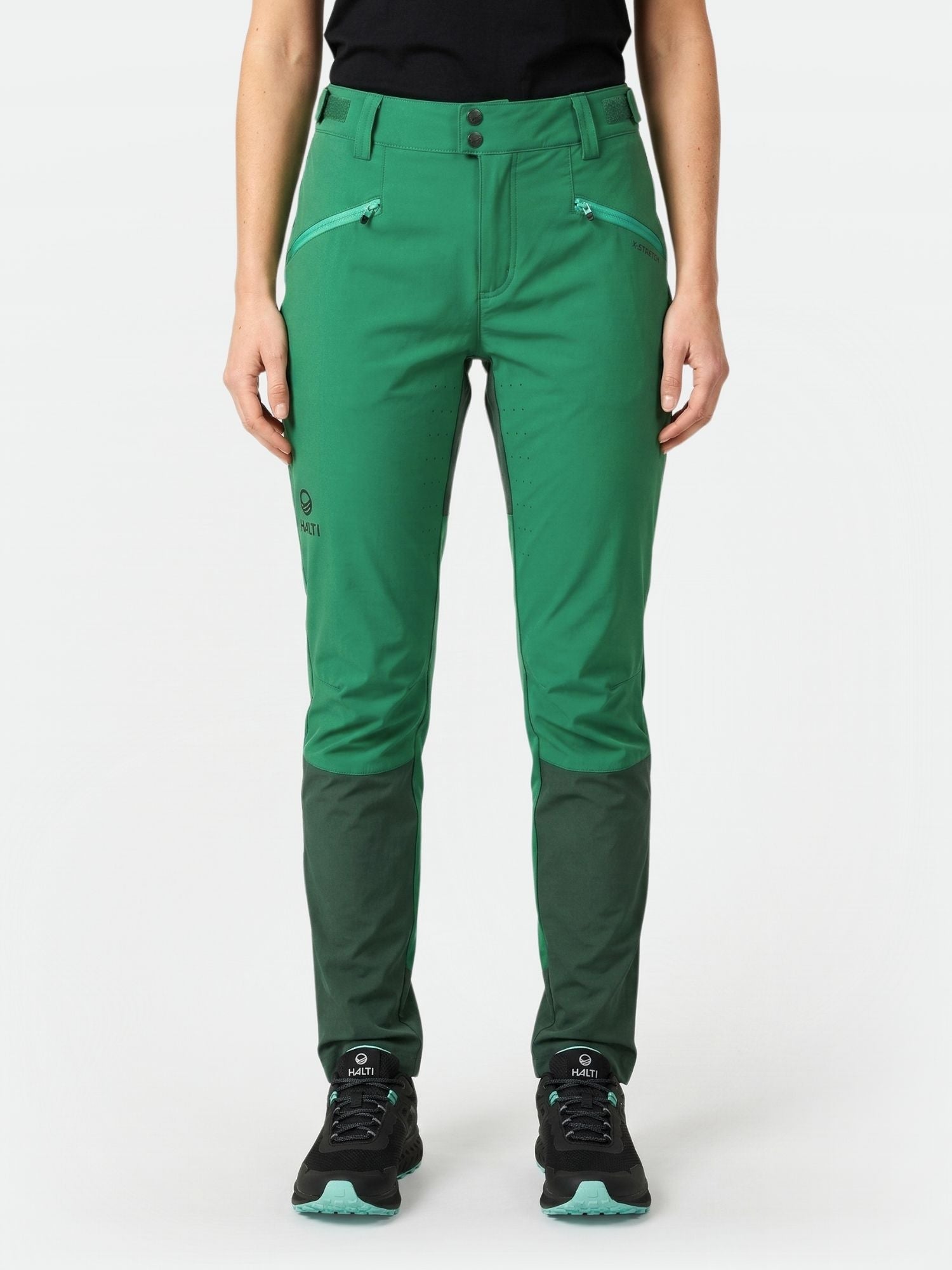 A person wears Halti Tracks Women's X-stretch Pants with zipped pockets and black shoes, facing forward against a plain background.