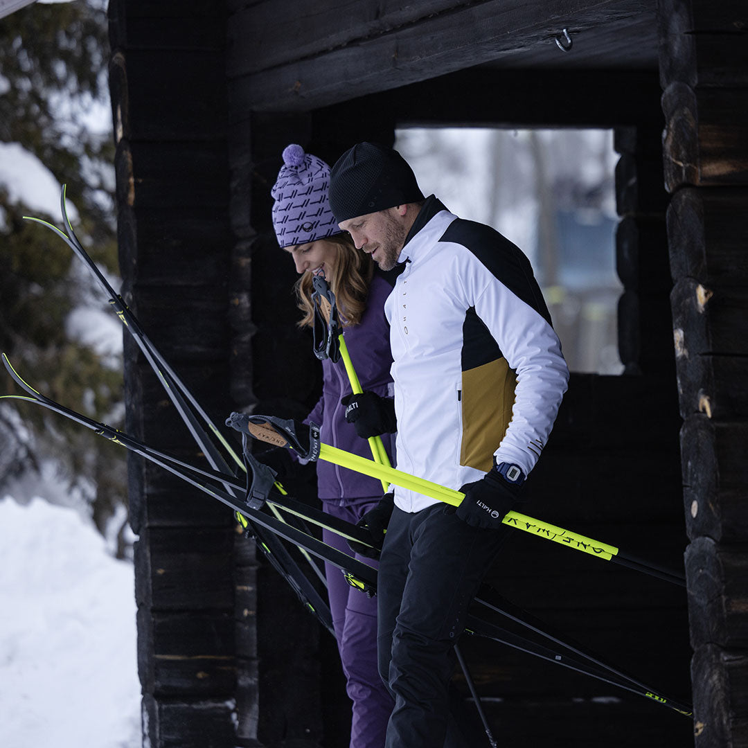 Two people in Halti Veloce XCT men’s ski jackets hold skis and poles by a snowy wooden cabin entrance.