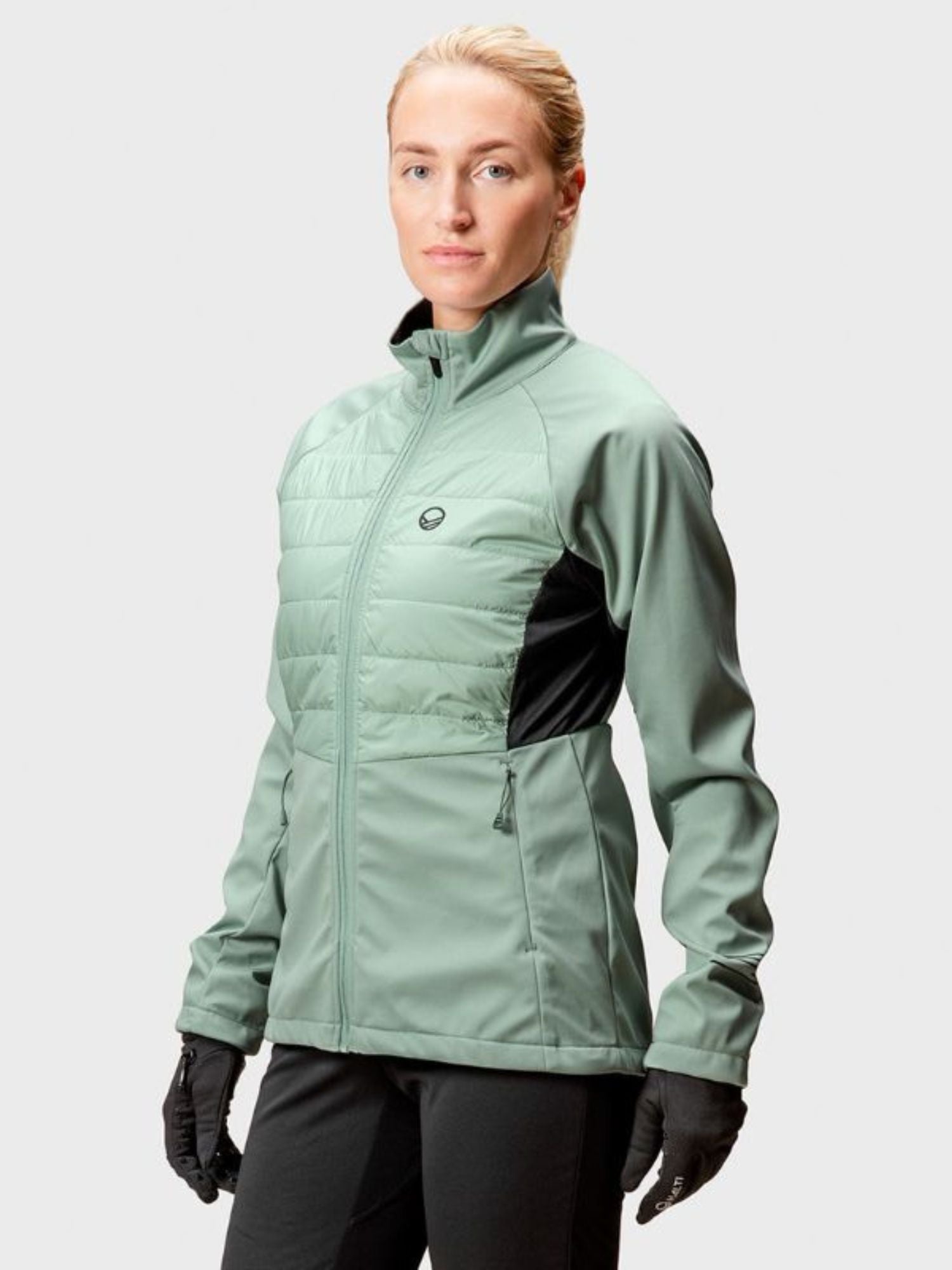 A person wears a Halti Vinha Hybrid XCT Jacket Women's in light green, black gloves, and pants against a plain light background.