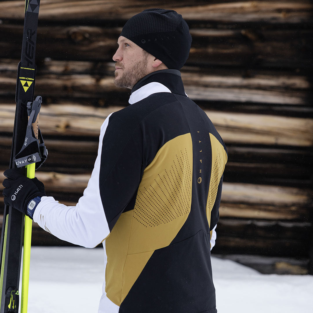 Wearing the Halti Veloce XCT Jacket, a skier stands on snow by a log wall, facing away from the camera.