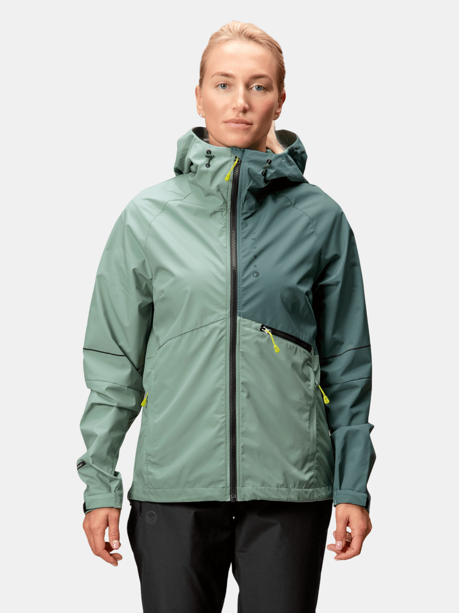 A woman in the Halti Shelter Stormwall 3L Shell Jacket—a two-tone green women’s shell with yellow zippers, ideal for winter.