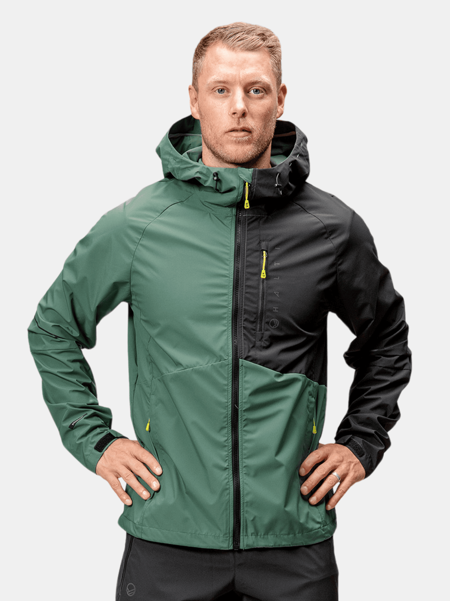 A man wears the Halti Shelter Stormwall 3L mens jacket in green and black, standing hands on hips against a plain backdrop.