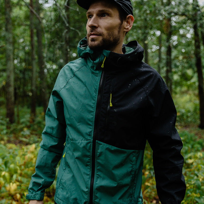 A man in a Halti Shelter Stormwall 3L Jacket stands in a forest, looking sideways with trees behind him.