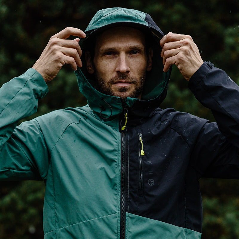 A man wears the Halti Shelter Stormwall 3L Jacket, holding up its hood outdoors.