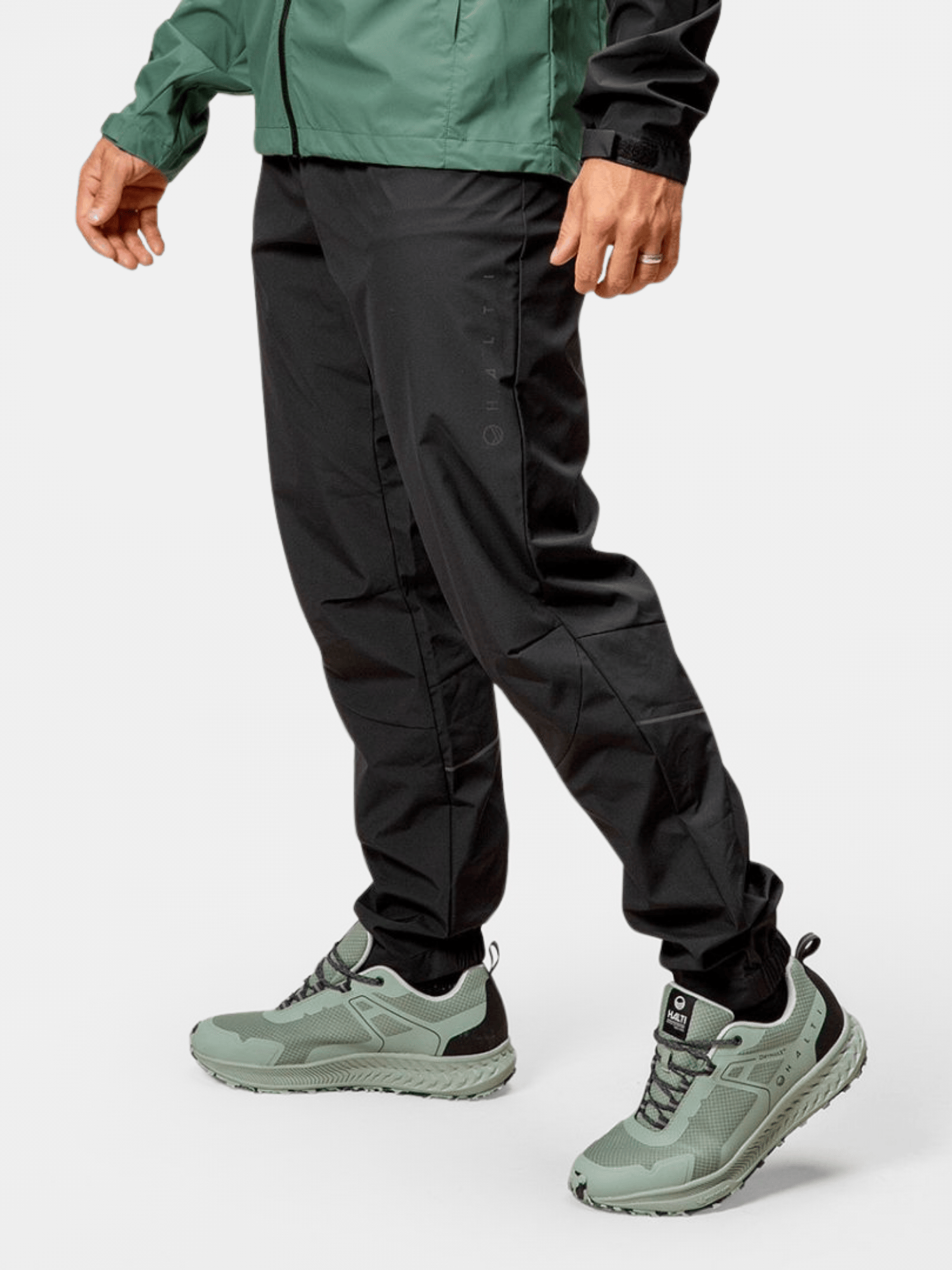 A person in Halti Shelter Stormwall 3L Pants, a green jacket, and light green running shoes stands against a plain background.