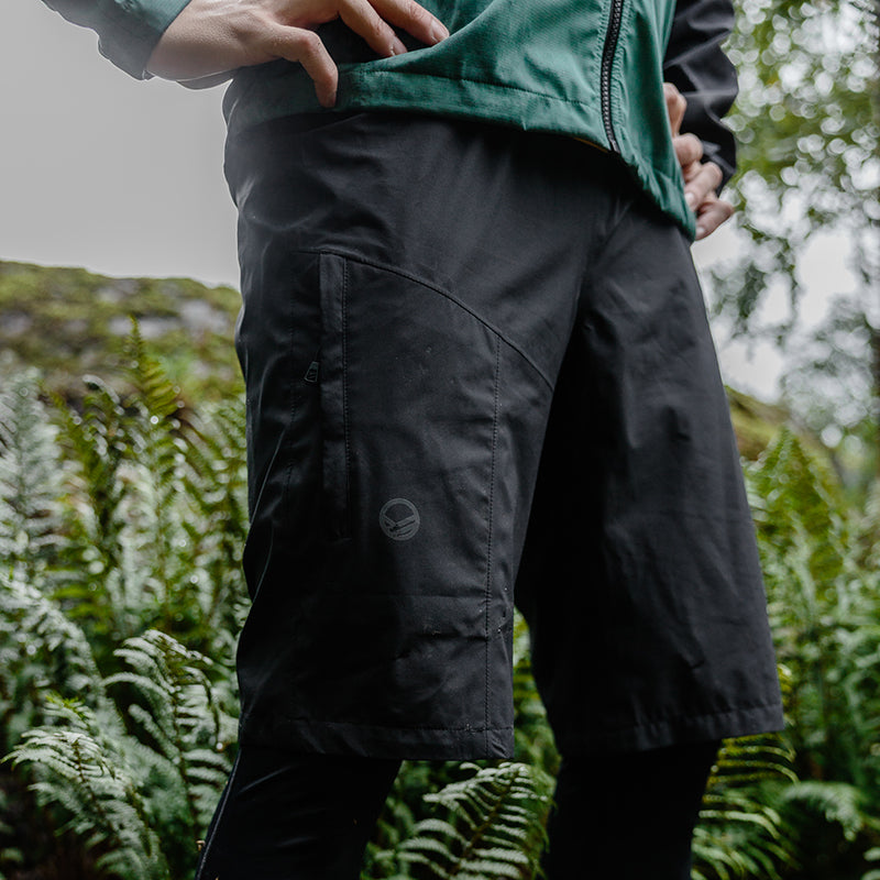 A person in Halti Shelter DrymaxX Unisex Shorts and a green jacket stands outdoors among lush green ferns.