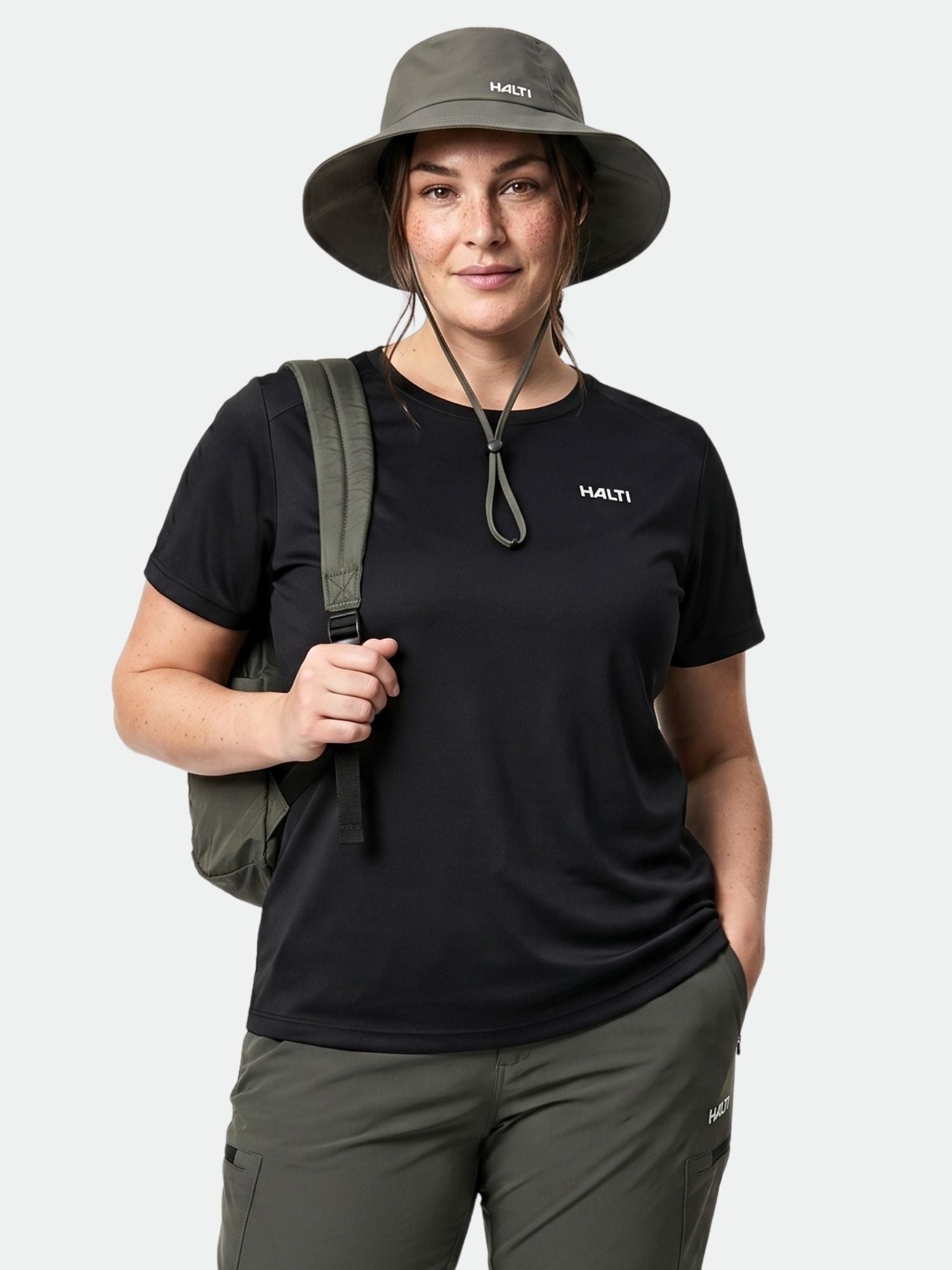 A woman in a Halti Salves T-shirt Women's Plus, olive pants, and a backpack stands against a plain background.