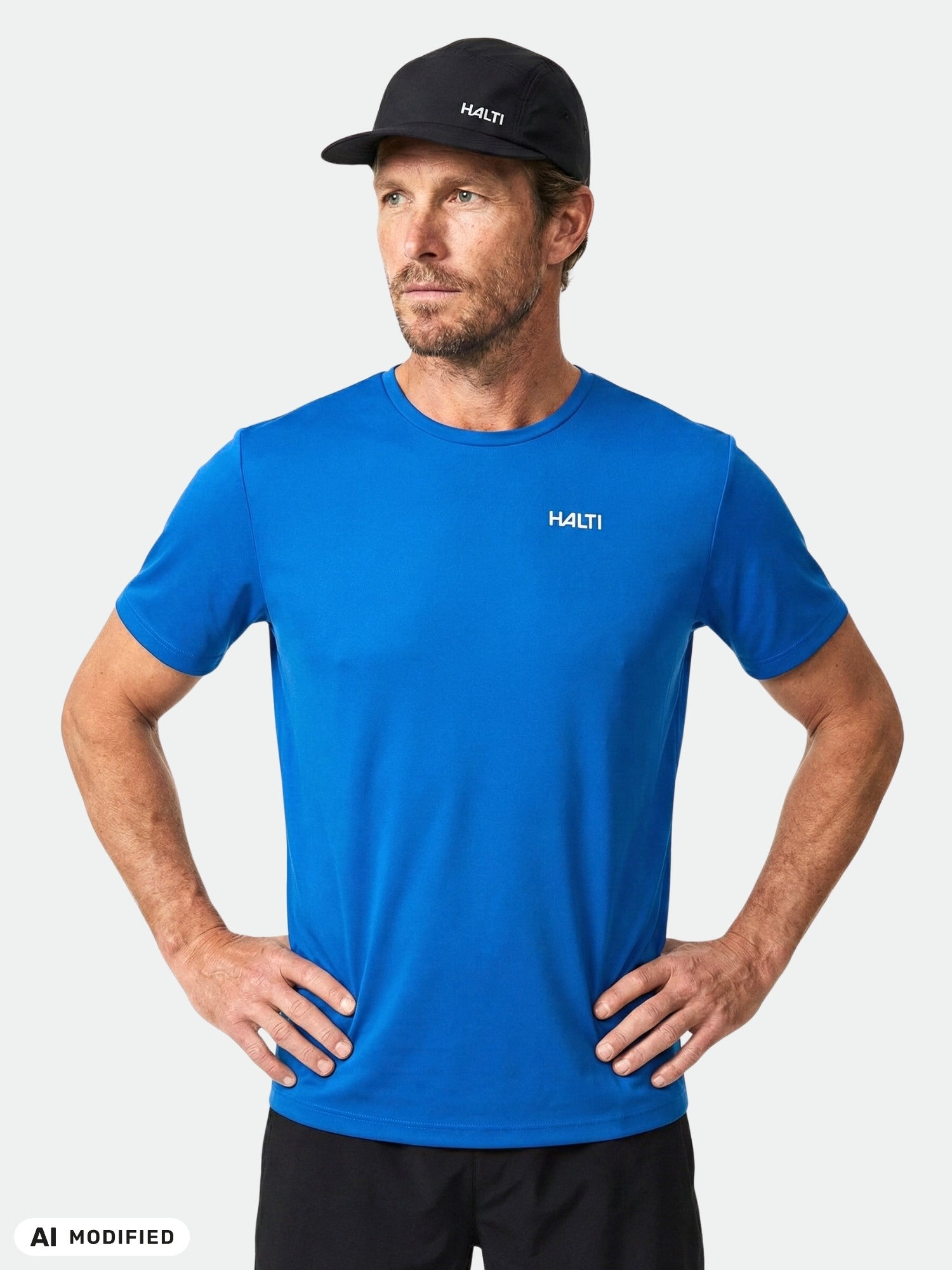 Man in a blue Halti Salves T-shirt and black cap stands with hands on hips against a plain light background.
