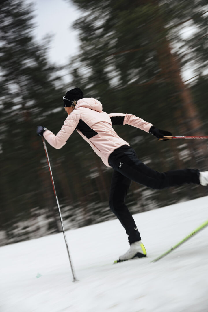 A woman in Halti Hanki 2 Hybrid XCT Pants cross-country skis on snow with trees in the background.