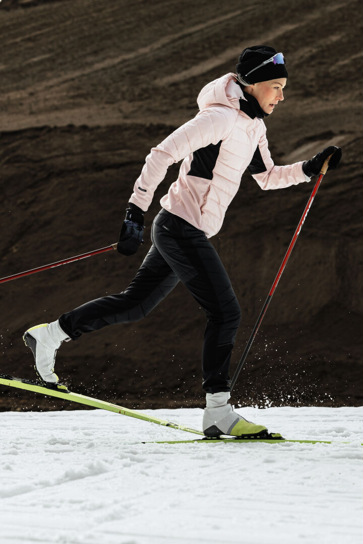 A woman cross-country skis on snow in Halti Hanki 2 Hybrid XCT Pants, holding ski poles against a rocky backdrop.