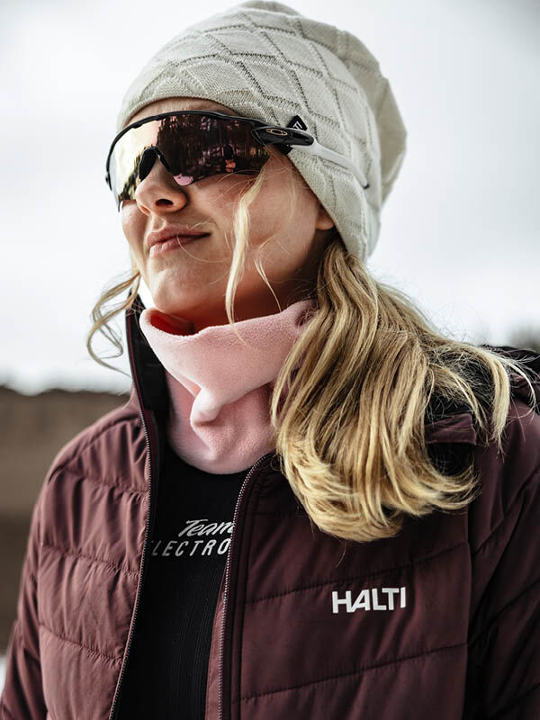 Woman outdoors in a Halti Crostie Merino Beanie, sunglasses, pink scarf, and maroon Halti jacket.