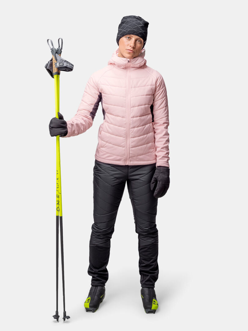 Woman in Halti Hanki 2 Hybrid XCT Jacket, black pants, gloves, hat, holding ski poles and standing on skis.
