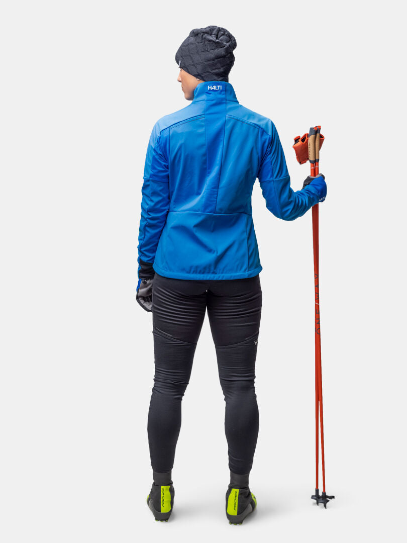 Woman in Halti Framover Hybrid XCT Jacket and black leggings, holding red ski poles, faces away from the camera.