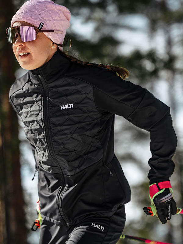 Woman cross-country skiing in a pink beanie and Halti Framover Hybrid XCT Jacket, forest in the background.