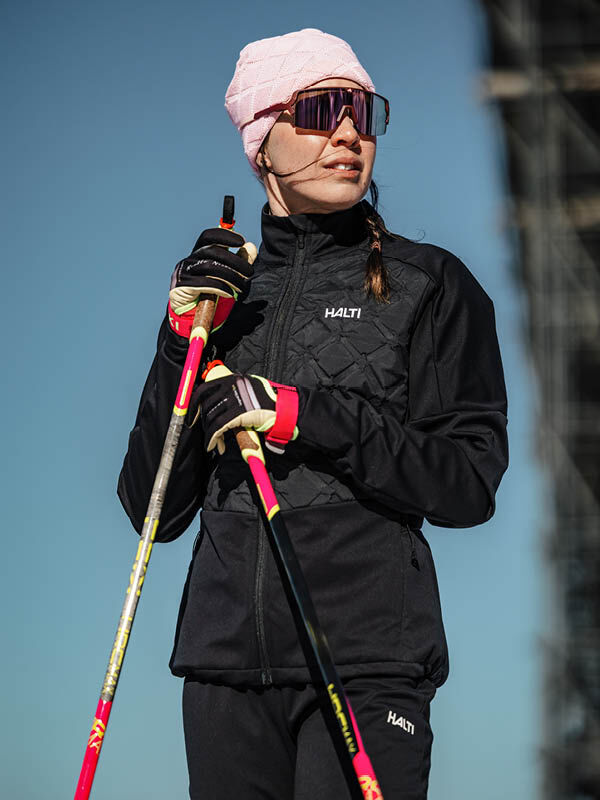 Wearing a Halti Framover Hybrid XCT Jacket, a woman in a pink beanie and sunglasses holds ski poles outdoors.