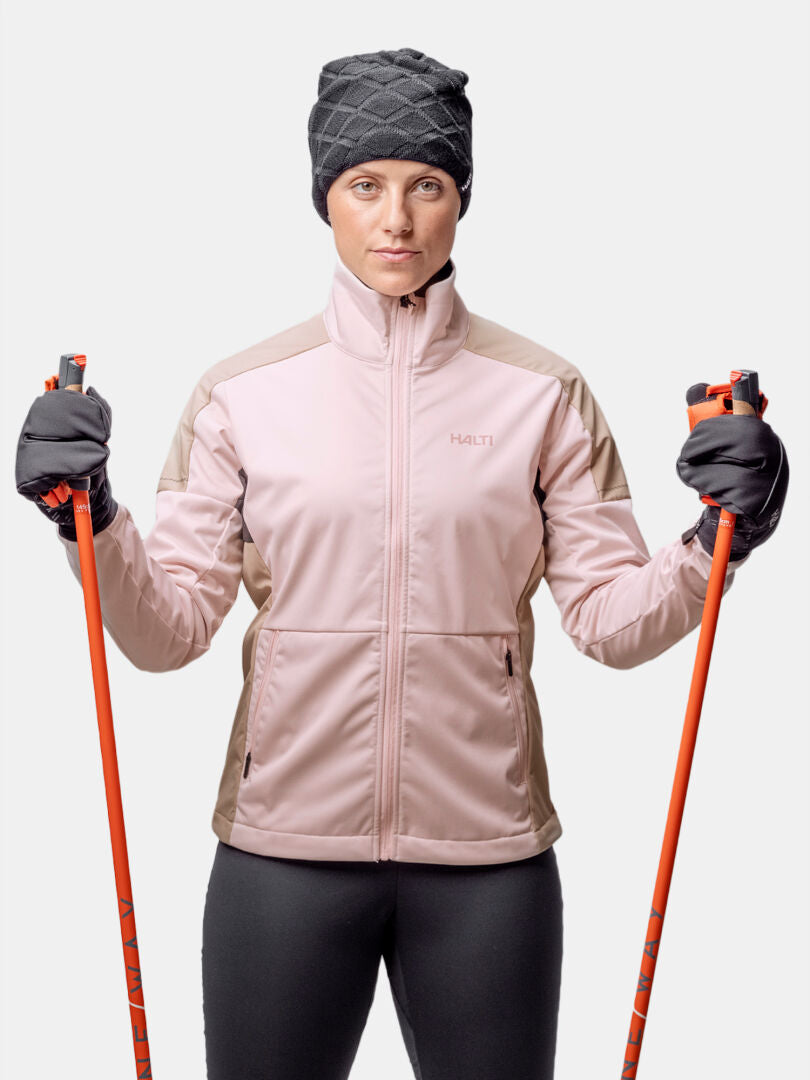 A woman in the Halti Huuru XC Set Women's holds two orange ski poles, ready for cross-country skiing on white background.