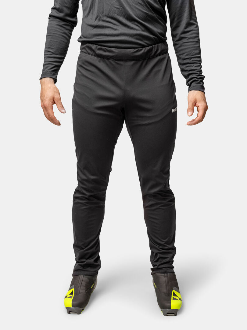 A person stands wearing Halti Framover XCT Pants Men's, a black long-sleeve shirt, and black shoes with yellow accents.