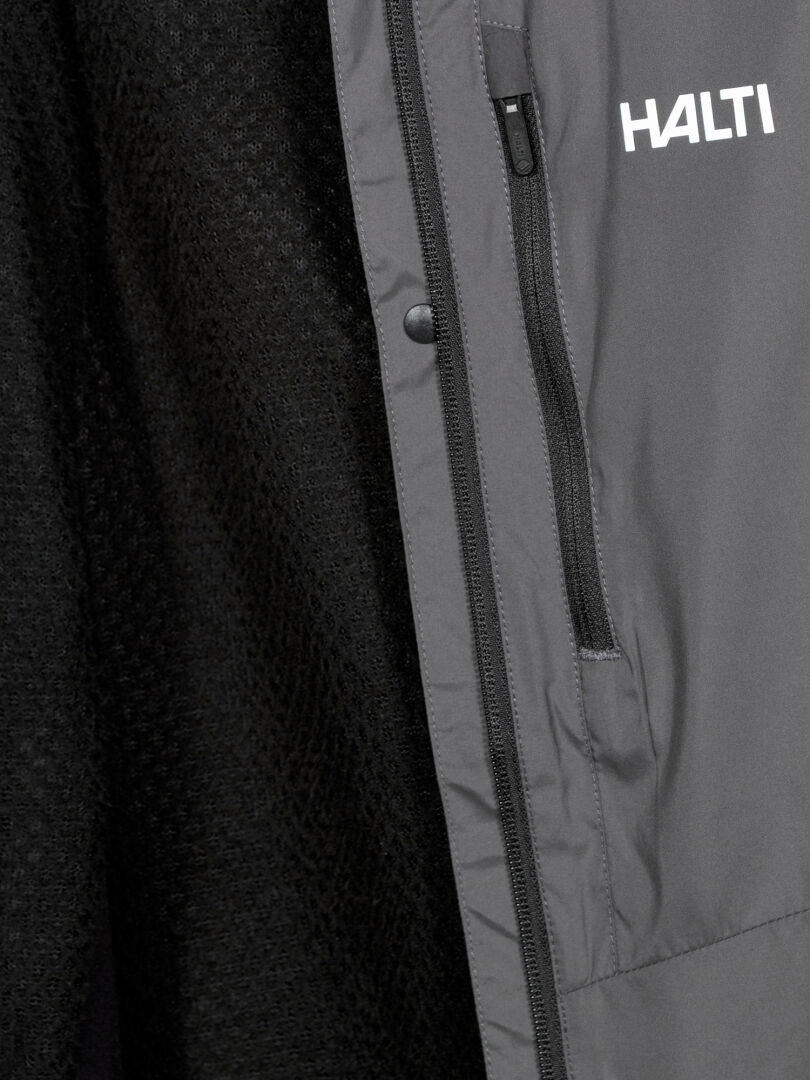 Close-up of a grey Halti Framover XCT Unisex skiing vest with zip pocket and logo, next to black textured fabric.