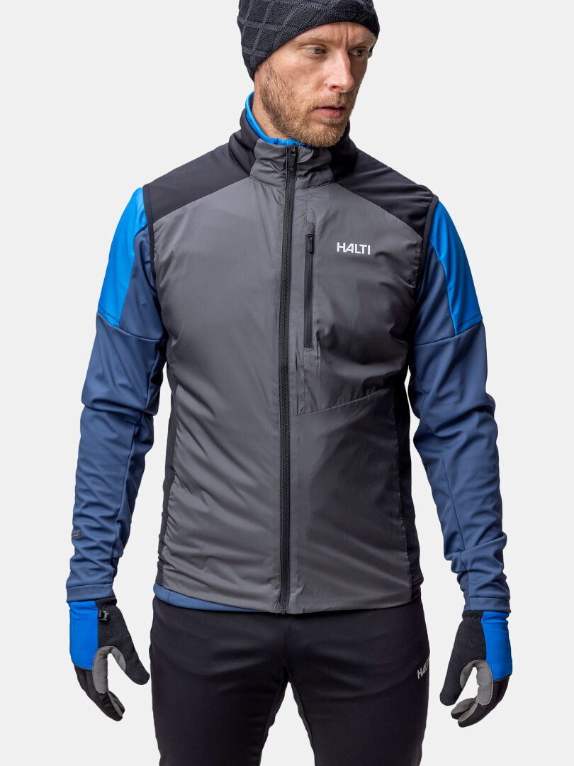 A man wears the Halti Framover XCT Vest Unisex in gray and blue, with black gloves and hat, against a plain background.