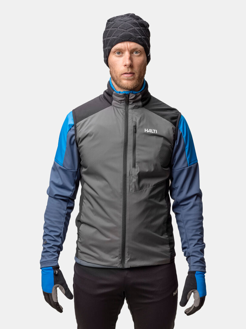A man wears a Halti Framover XCT Vest Unisex in gray/blue, black gloves, hat, and pants against a white background.