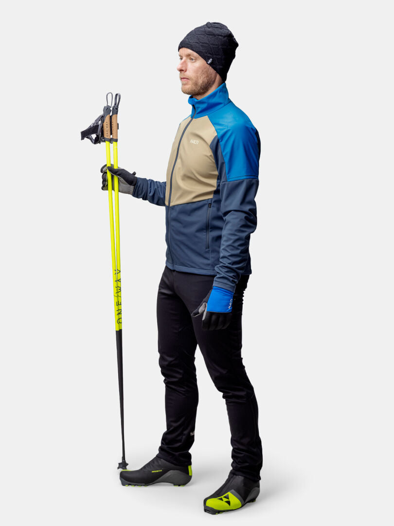 A man in a Halti Framover XCT Jacket holds yellow ski poles, wearing gloves, a beanie, and athletic shoes.