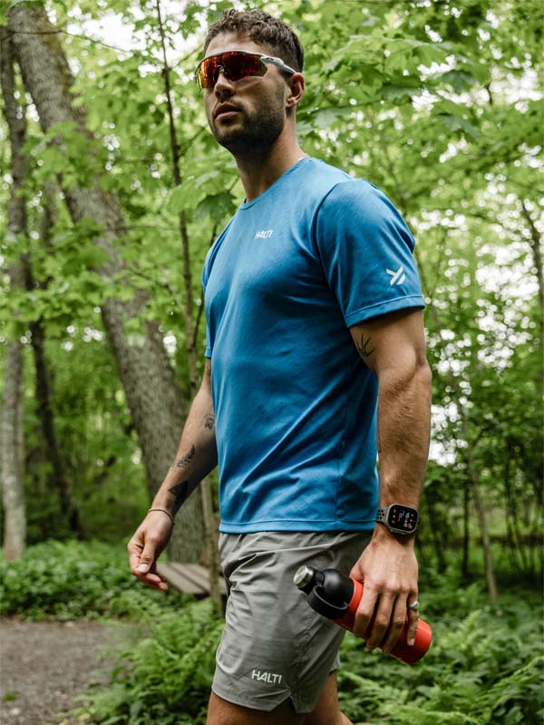 Wearing a Halti Oulanka T-shirt and grey shorts, a man walks in the forest with a water bottle, sunglasses, and smartwatch.