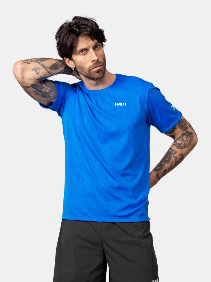 A man in a blue Halti Oulanka T-shirt and black shorts stands with one arm raised, looking at the camera.