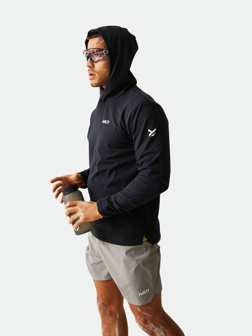 A man in the Halti Kaltio Training Hoodie holds a water bottle, wearing gray shorts and sunglasses against a plain background.