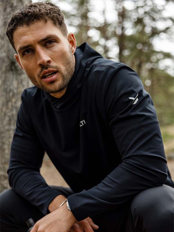 A man in a Halti Kaltio Training Hoodie sits outdoors in a wooded area, looking slightly to the side.