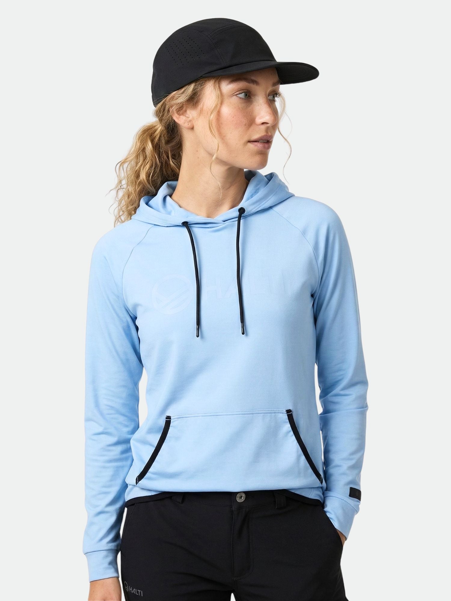 Woman wearing Halti Veidnes Women's hoodie in light blue, black pants, and cap, looking left against plain background.