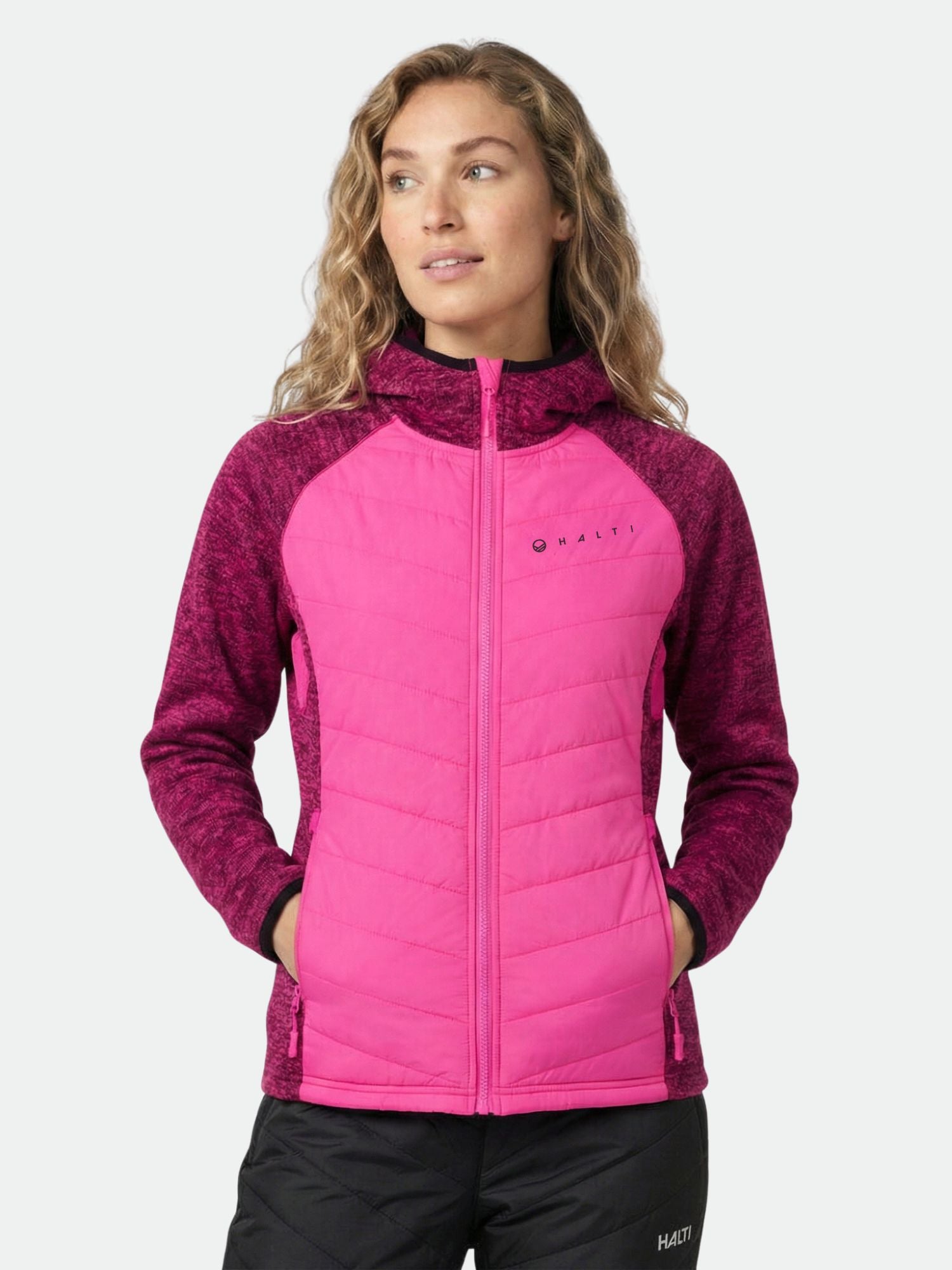 Woman in Halti Streams Hybrid Layer Jacket, bright pink and maroon, hands in pockets, looking to the side.