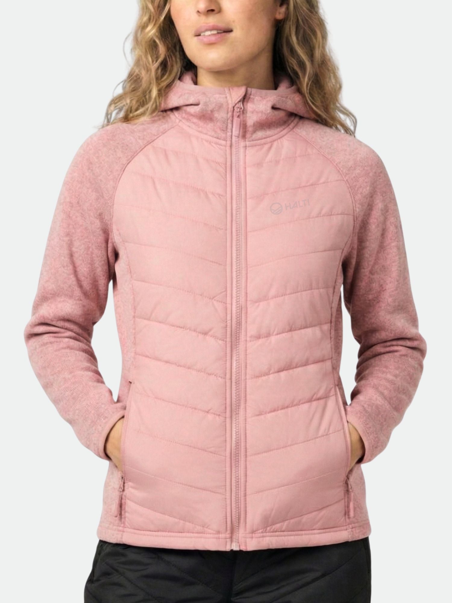 A woman wears the Halti Streams Hybrid Layer Jacket, light pink with front zipper and pockets, against a plain background.