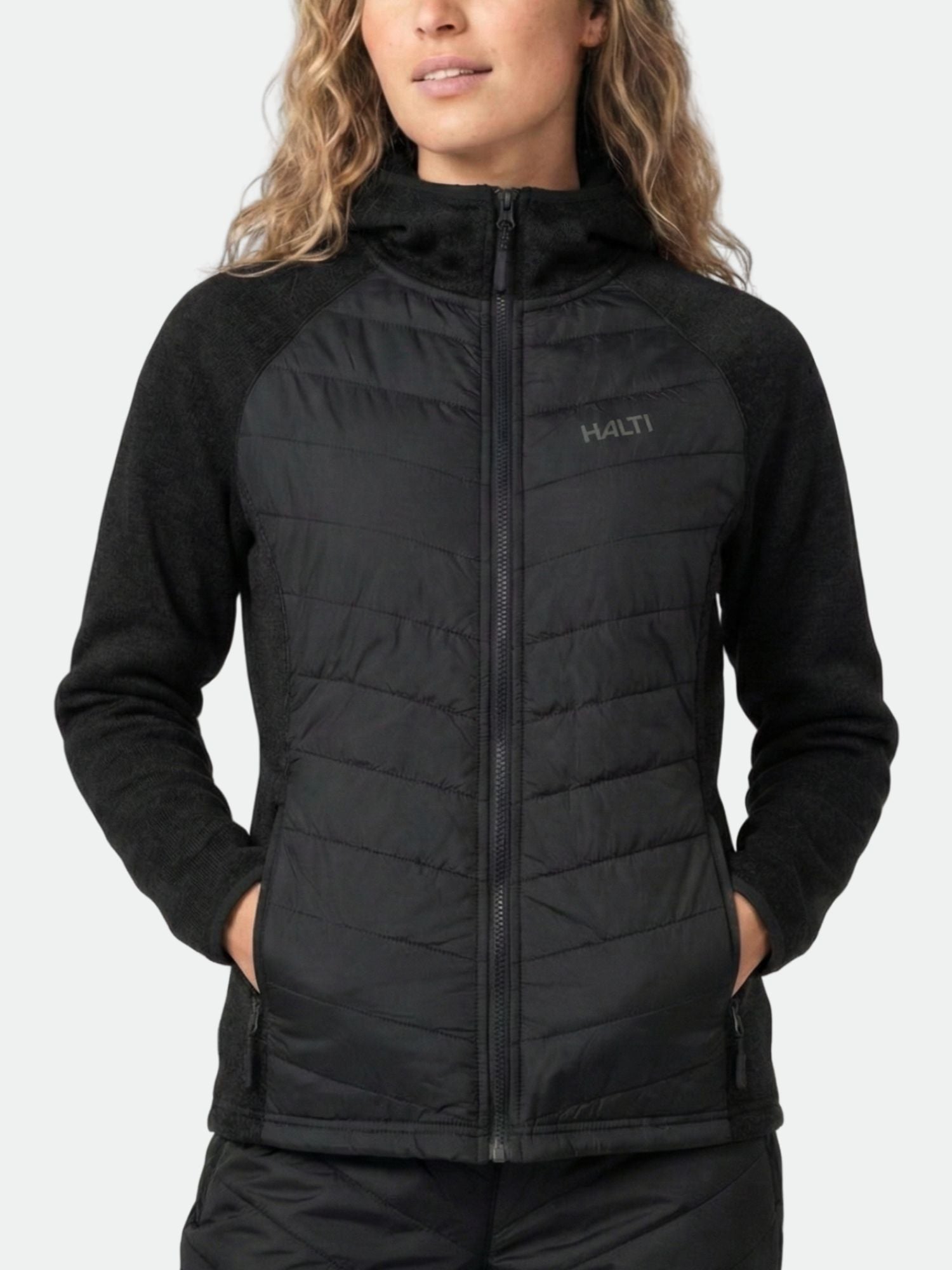 A woman in a black Halti Streams Hybrid Layer Jacket stands facing forward, hands in pockets, against a plain background.
