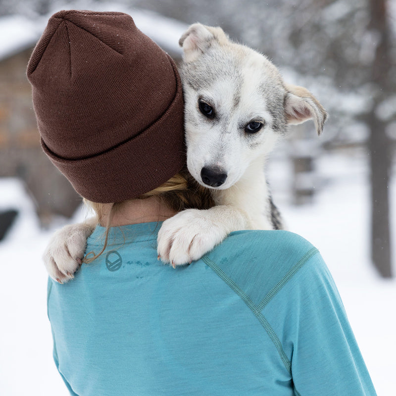 A woman in a Halti Pihka II Merino Baselayer shirt holds a dog over her shoulder in the snow.