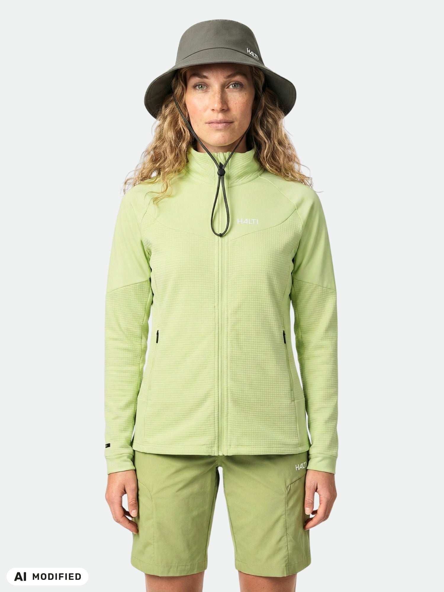 Woman wears Halti HeatGrid Layer Jacket and matching shorts with an olive bucket hat, set against a plain background.