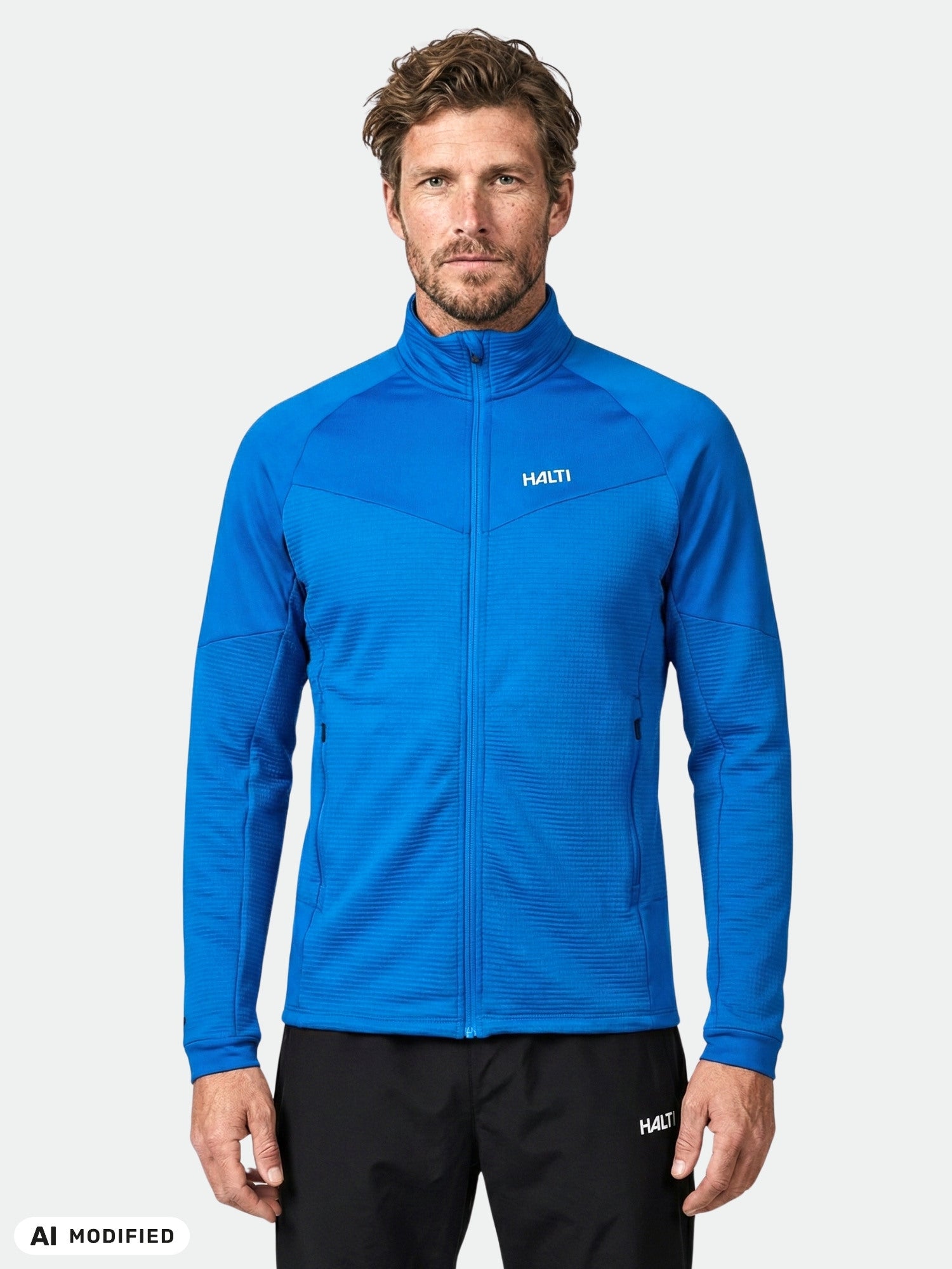 Man in Halti HeatGrid Layer Jacket, bright blue waffle-knit, with black pants, stands before a light background.