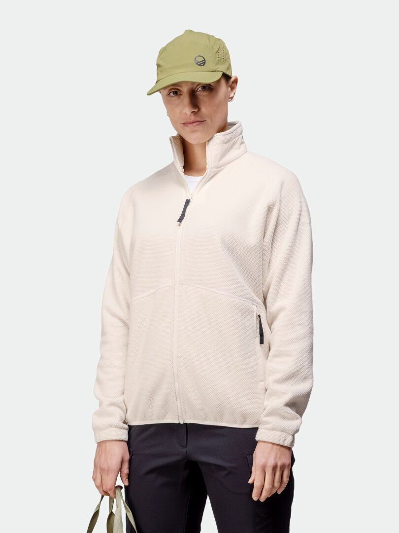 A woman in a Halti Soolo Layer Jacket in light beige, black pants, and a green cap stands against a plain background.