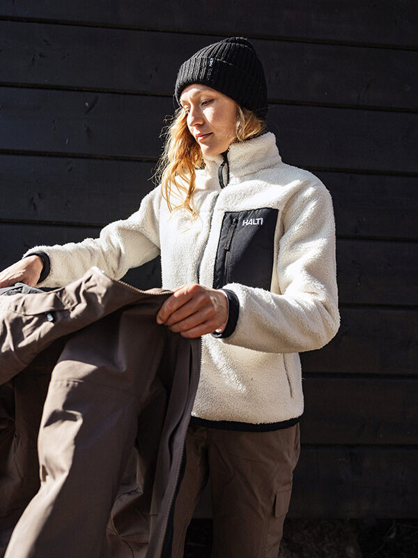 A woman wearing a Halti Lumista Polar Fleece Jacket and black beanie holds a brown coat by a dark wooden wall.