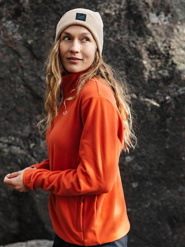 A woman in a Halti Rockmoon Fleece Jacket and beige beanie stands by a rock outdoors, looking to the side.