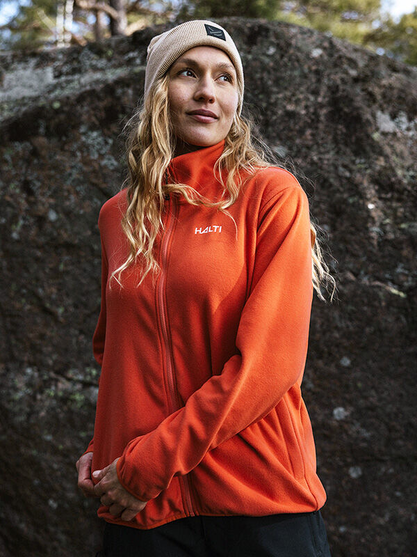 A woman in a Halti Rockmoon Fleece Jacket and beige beanie stands outdoors by a large rock.