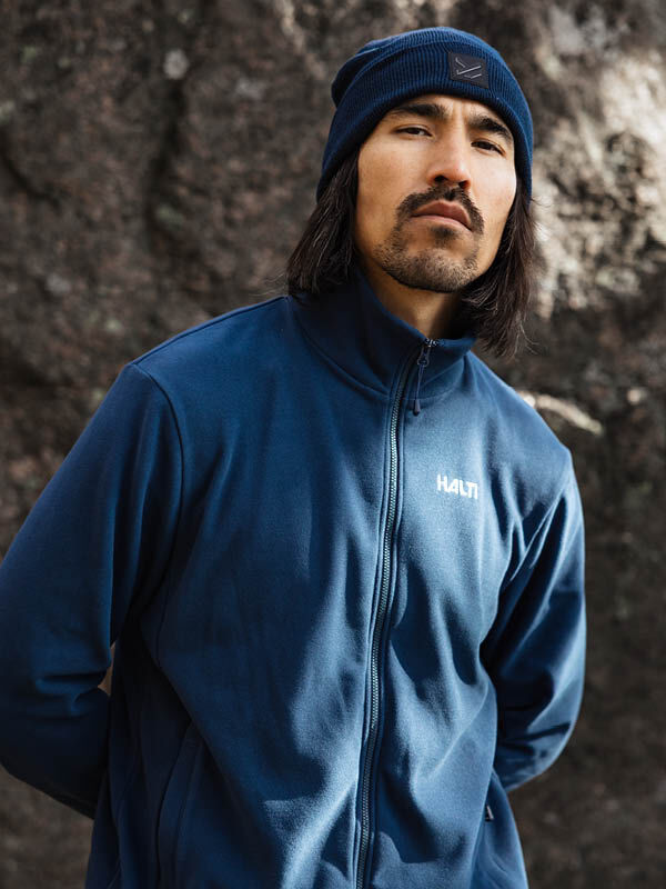 A man wears the Halti Rockmoon Fleece Jacket Men's in blue and a beanie, standing outdoors by rocks.