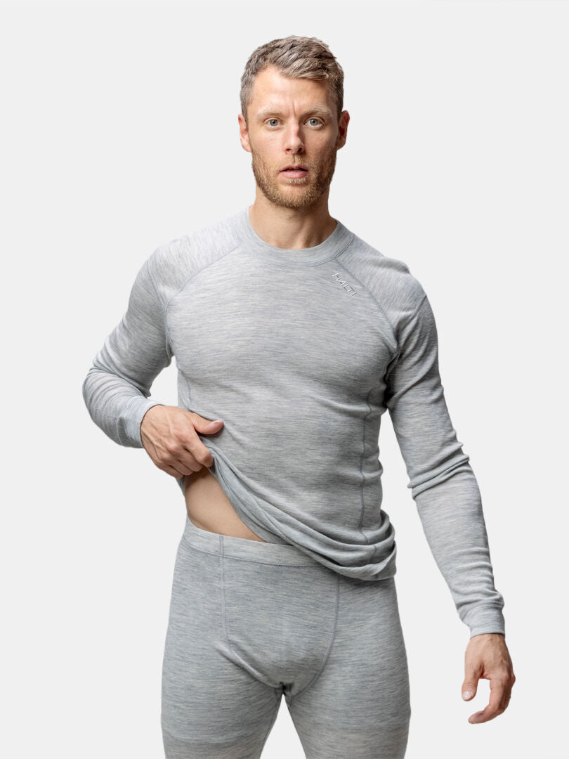 Man wears Halti Hossa II Merino Base Layer Set in light gray, lifting shirt slightly against a plain background.