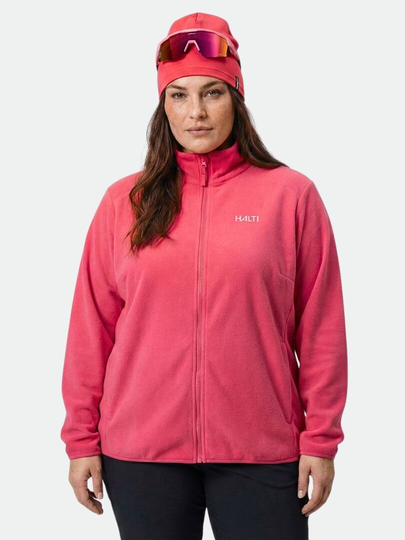 A person in a Halti Rockmoon Fleece Jacket Women’s Plus, pink beanie, and goggles stands by a plain light background.
