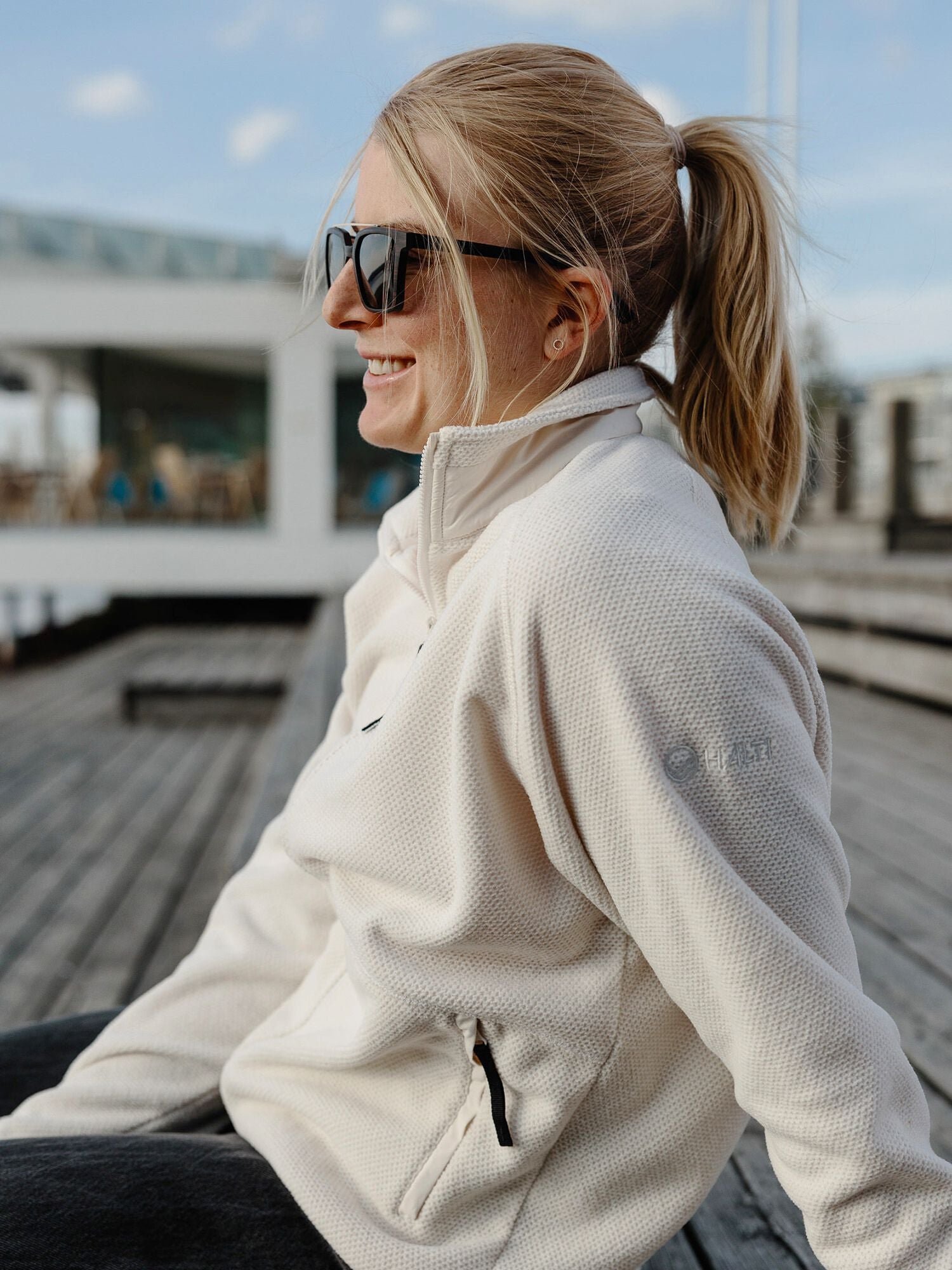 A woman smiles outdoors in the Halti Soolo Layer Jacket Womens, featuring zippered pockets and stylish sunglasses.
