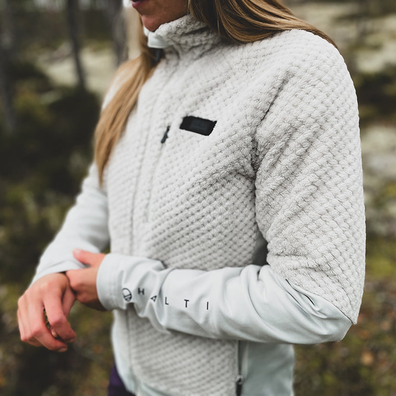 Woman outdoors in a textured light gray Halti Forceful Womens Layer Jacket, adjusting sleeve; Halti logo on forearm.
