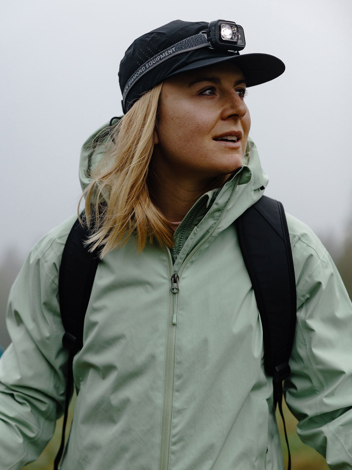 A person in a Halti Staala DX Shell Jacket Womens, black backpack, and headlamp looks sideways outdoors on a cloudy day.