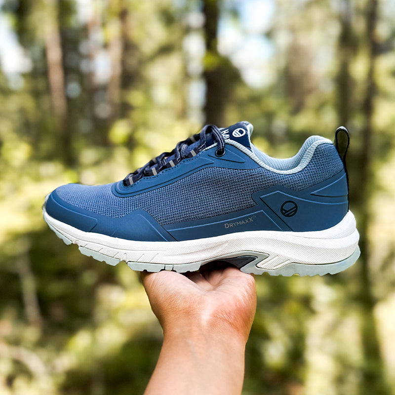 A hand holds a Halti Fara Low 2 DrymaxX Outdoor Shoe Womens in blue with GripmaxX sole against a green forest background.
