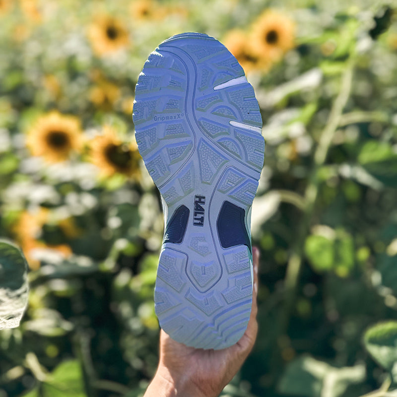 A Halti Fara Low 2 DrymaxX outdoor shoe is held up close, with blurred sunflowers and greenery in the background.