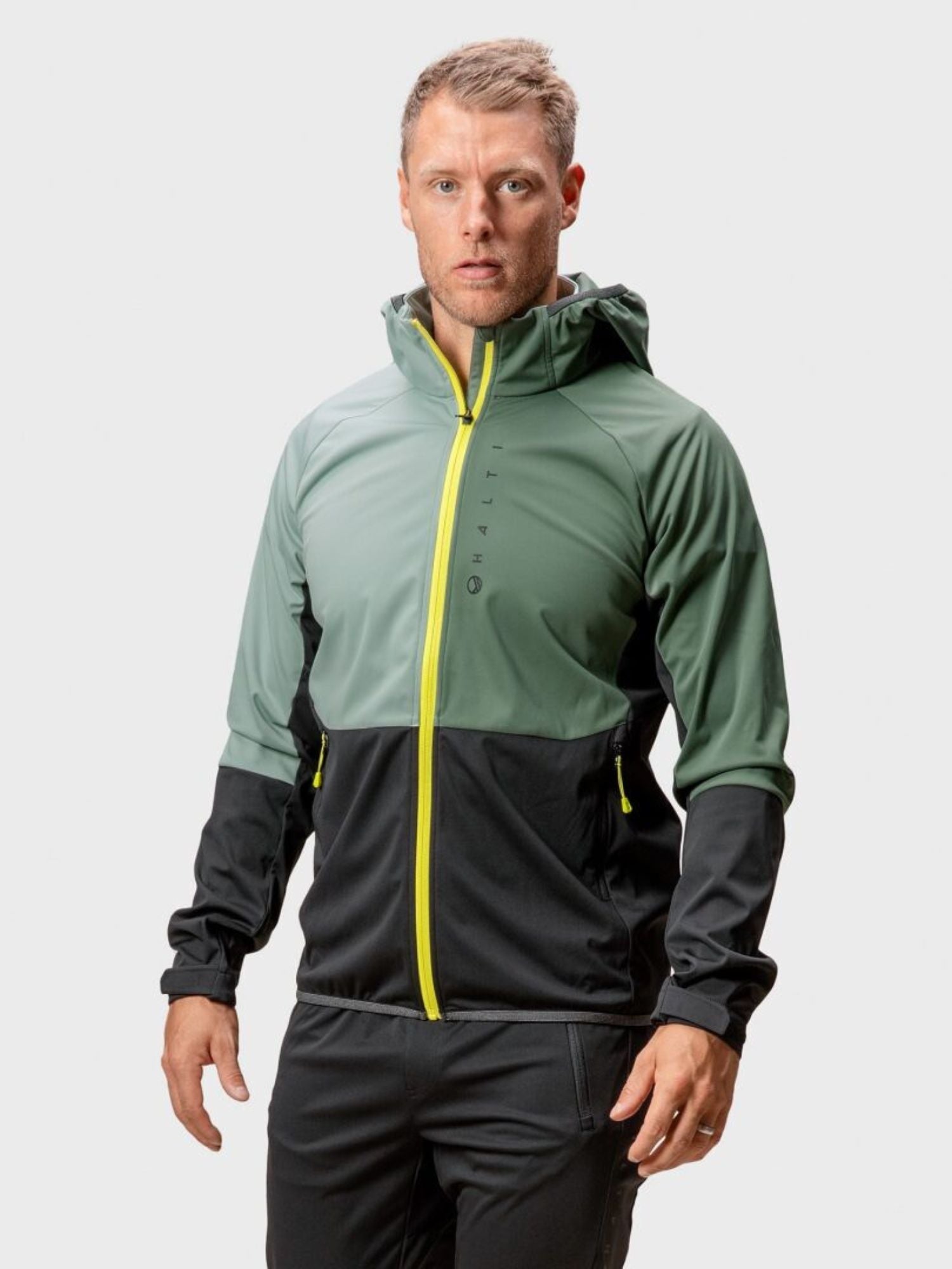 A man wears a Halti Exhale Stormwall Jacket Men's with a yellow zipper and reflective details against a plain background.